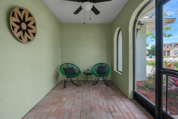 A small, enclosed porch area with two green chairs, a ceiling fan, and a decorative wall piece.