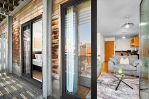 Et moderne interiør med en træfacade, en balkon med en glasdør, et opholdsområde med sofa og sofabord, samt et køkken der kan anes gennem glasset.