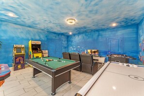 Una mesa de billar, una mesa de fútbolín y máquinas de videojuegos en una sala con paredes y techo azules.