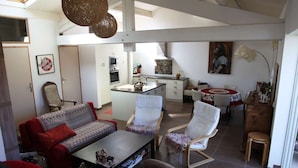 A spacious living area with a sofa, chairs, and a coffee table. A kitchen is visible in the background with a dining table and chairs. - Apartment with terrace overlooking the lake, a stone's throw from the port (Mèze)