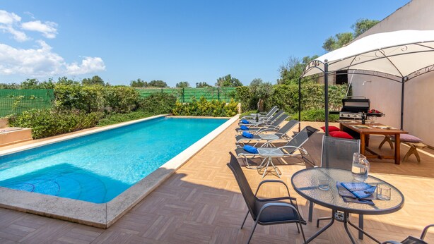 Une piscine entourée de verdure, avec des chaises longues et une table, sous un ciel dégagé.