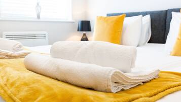 A neatly made bed with white towels and a yellow blanket, a bedside lamp, and a window with blinds.