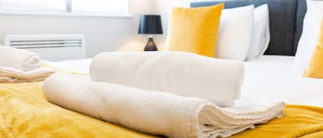 Una cama bien hecha con toallas blancas y una manta amarilla, una lámpara de noche y una ventana con persianas.