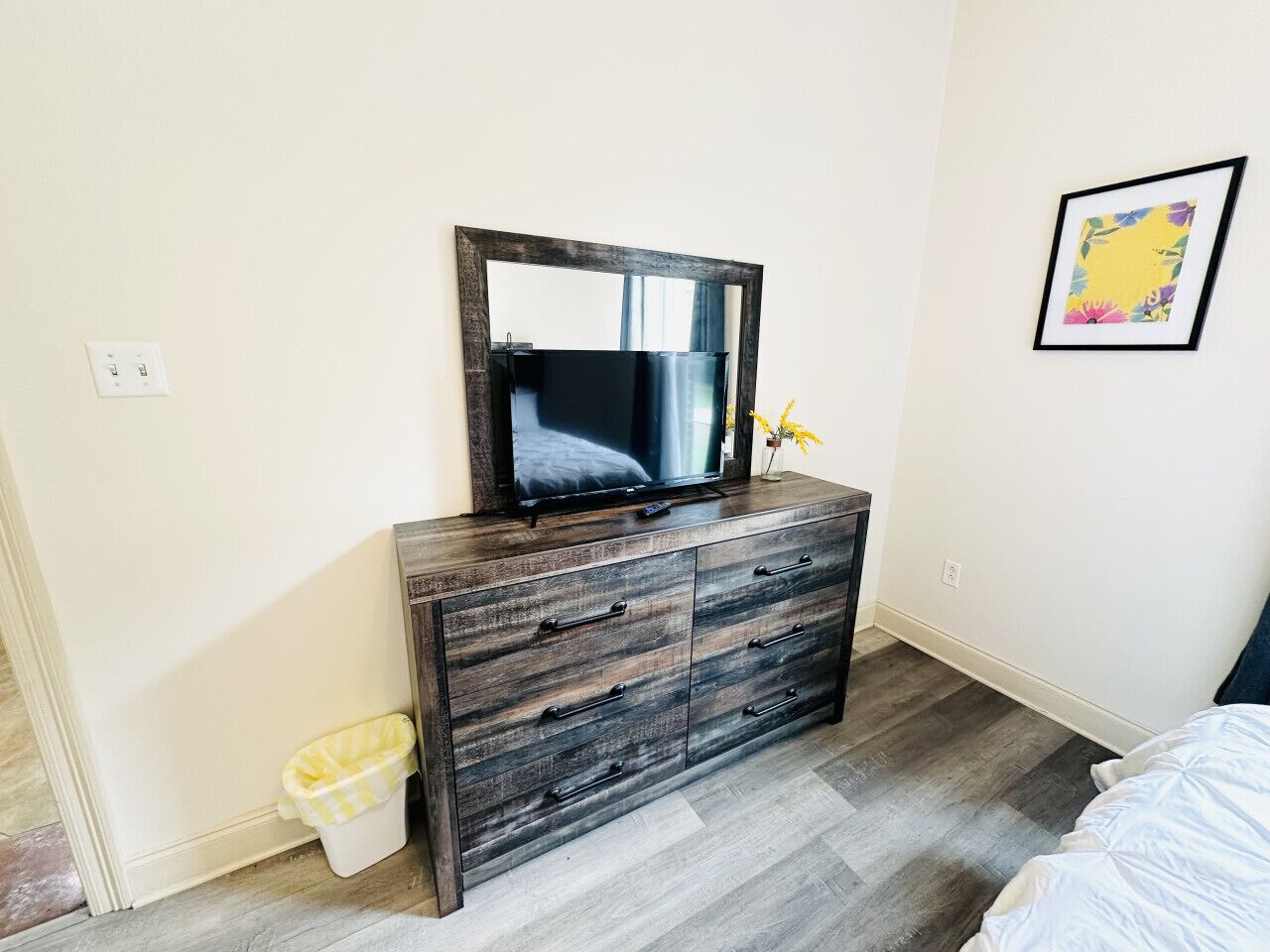 A bedroom with a wooden dresser, a mirror, a bed, and a small trash bin.