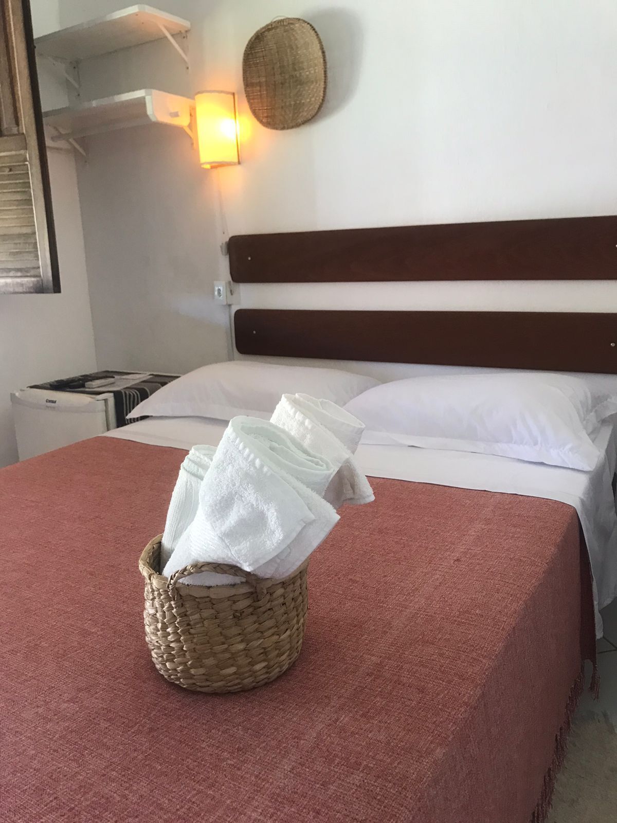 Un lit bien fait, avec du linge de lit blanc, une tête de lit marron et un panier en osier contenant des serviettes pliées, posé sur une couverture rouge.