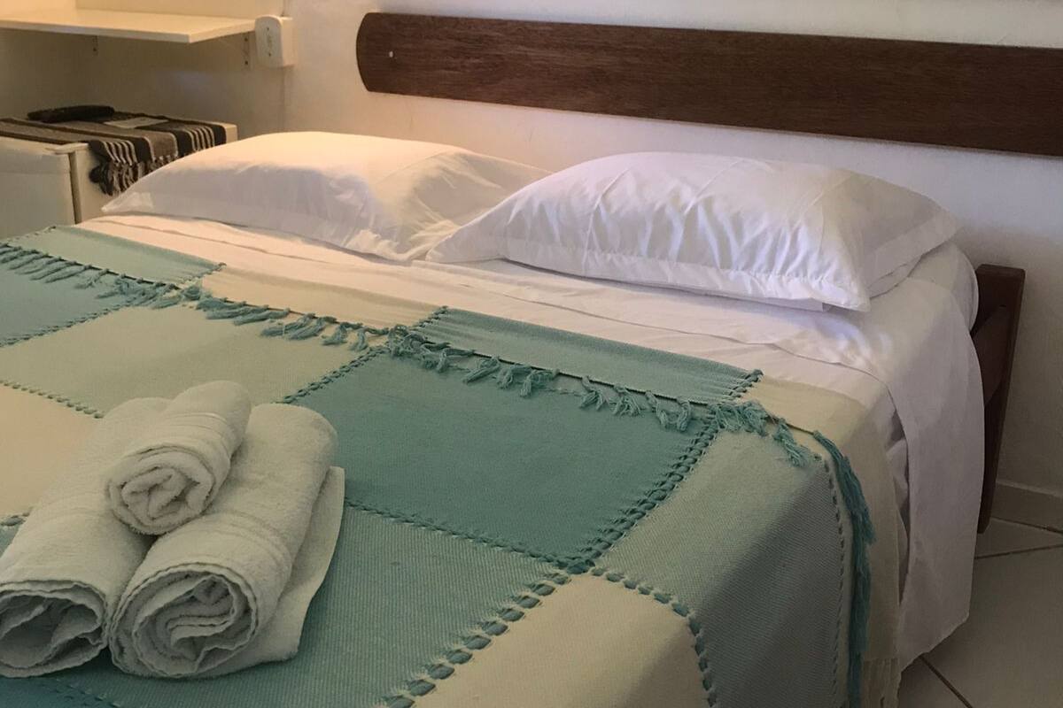 Una cama bien hecha con una colcha verde aguamarina y blanca, dos toallas blancas enrolladas y un cabecero de madera.