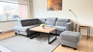 A modern living room with a grey sectional sofa, a wooden coffee table, and a small grey armchair. A painting hangs on the wall, and a floor lamp is visible. - North Sea Residence App 23 Westerland (Westerland)