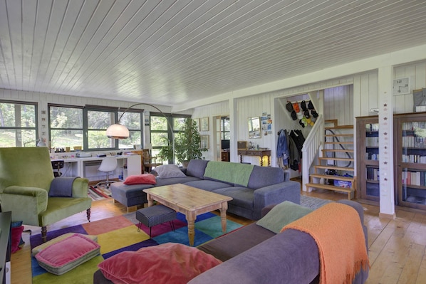 Un salon spacieux avec un grand canapé, une table basse en bois et un tapis coloré. On y trouve un bureau avec une chaise et une lampe, ainsi qu’une bibliothèque remplie de livres.
