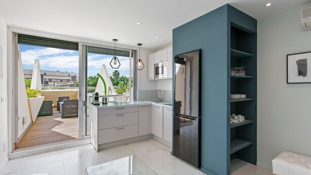 A modern kitchen with a large island, a built-in oven, and a refrigerator. There is a sliding glass door leading to an outdoor patio area with a wooden deck and outdoor furniture. - Naturist Village - Le Diamond X Héliopolis Luxe, Naturist Village (Agde)