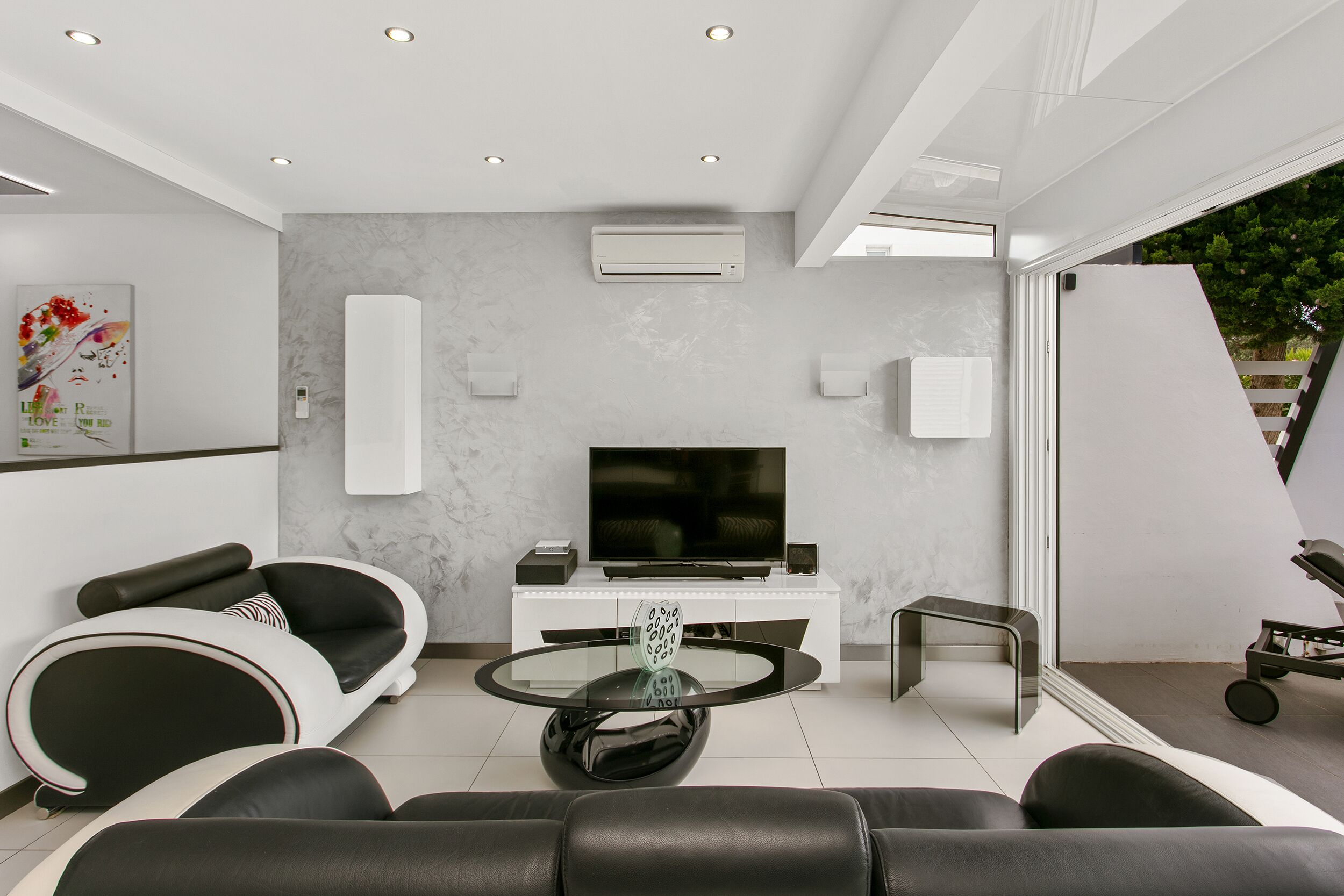 A modern living room with a flat-screen TV, a glass coffee table, and black leather sofas.