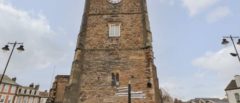 A historic stone tower with a clock, surrounded by a paved area with benches and street lamps.