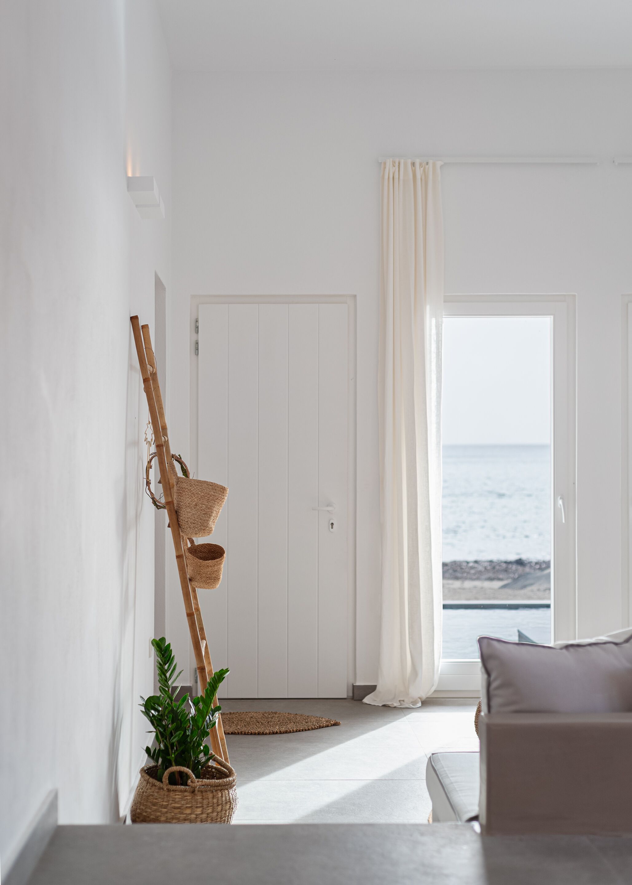A minimalist room with a view of the sea, featuring a white door, a plant, and a woven shelf.