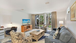 A living room with a sofa, armchair, coffee table, TV, and a desk with a lamp. - Villa 290 - updated condo with gorgeous ocean views (Jekyll Island)