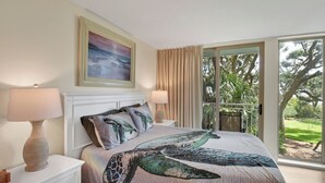 A bedroom with a large bed, two bedside lamps, a framed painting, and a sliding glass door leading to a balcony with a view of trees. - Villa 290 - updated condo with gorgeous ocean views (Jekyll Island)