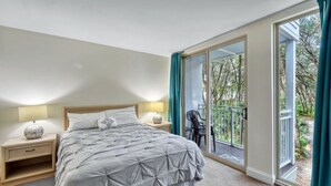 A bedroom with a large bed, two bedside tables, a nightstand, and a balcony with a chair. - Villa 452 - lovely condo just steps away from pool and beaches (Jekyll Island)