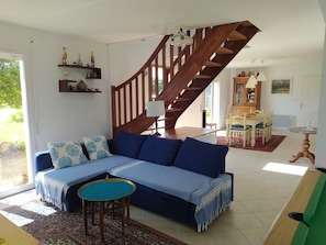 Un salon avec un canapé bleu, un escalier en bois, un coin repas avec une table et des chaises, et une bibliothèque.