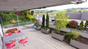 Un espace extérieur abrité, doté d’une table et de chaises, de jardinières contenant diverses plantes et offrant une vue sur le paysage environnant.