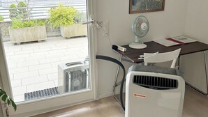 Une pièce comprenant un bureau, un ventilateur sur pied, un radiateur et une porte donnant sur un balcon agrémenté de plantes et offrant une vue sur le ciel.