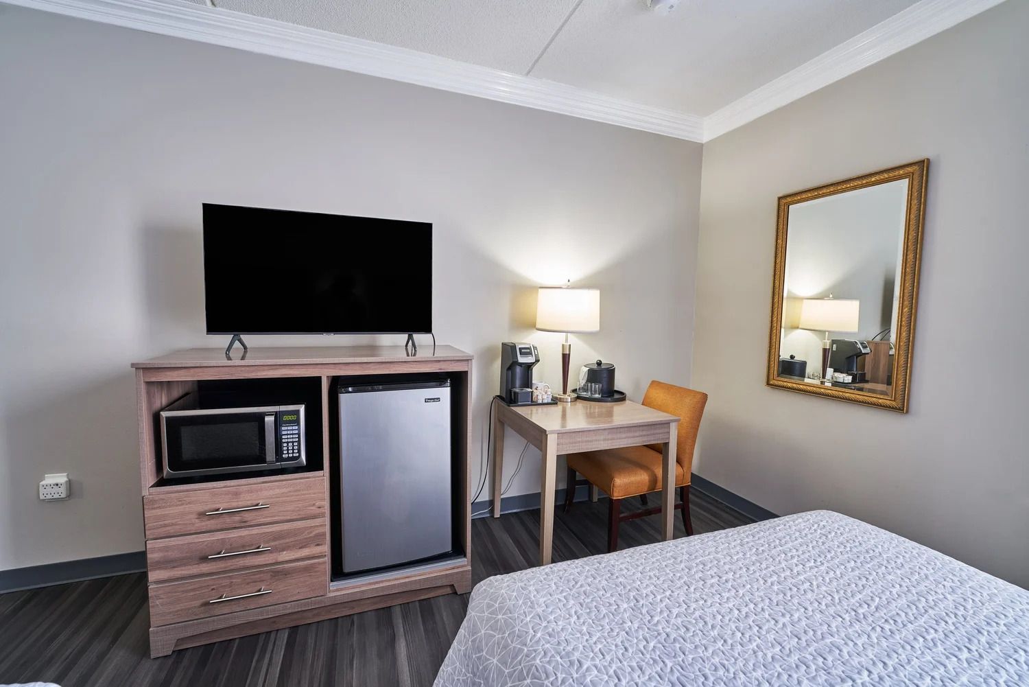 Ein Hotelzimmer mit einem Bett, einem Schreibtisch mit Stuhl, einem Fernseher, einer Mikrowelle, einem Kühlschrank, einer Lampe und einem Spiegel.