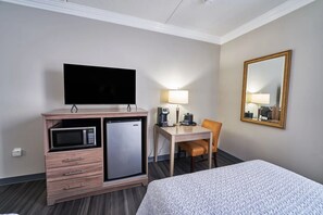 A hotel room with a bed, a desk with a chair, a television, a microwave, a refrigerator, a lamp, and a mirror.