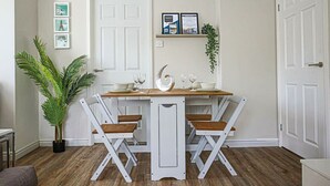 Uma sala de jantar com uma mesa e cadeiras brancas, uma planta em vaso e quadros nas paredes.