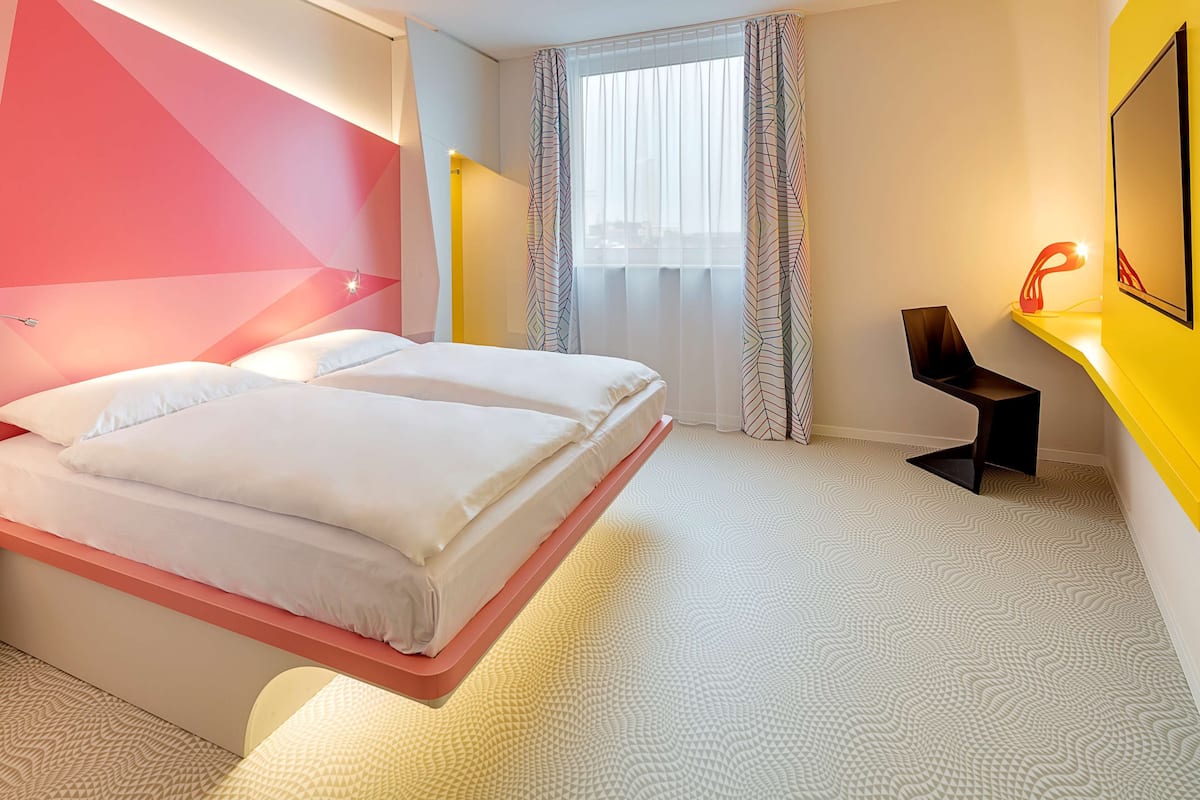 A modern hotel room with a bed, a desk, and a window with curtains.