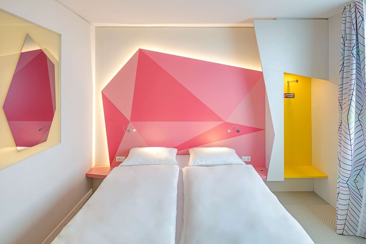 A modern hotel room with a geometric wall design, a bed with white linens, a small bedside table, and a shower area with a yellow accent wall.