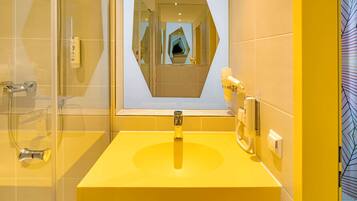 Un baño con un lavamanos amarillo, cabina de ducha con mampara de vidrio y un espejo con marco geométrico.