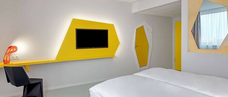 A modern hotel room with a bed, a desk with a flat-screen TV, and a mirror.