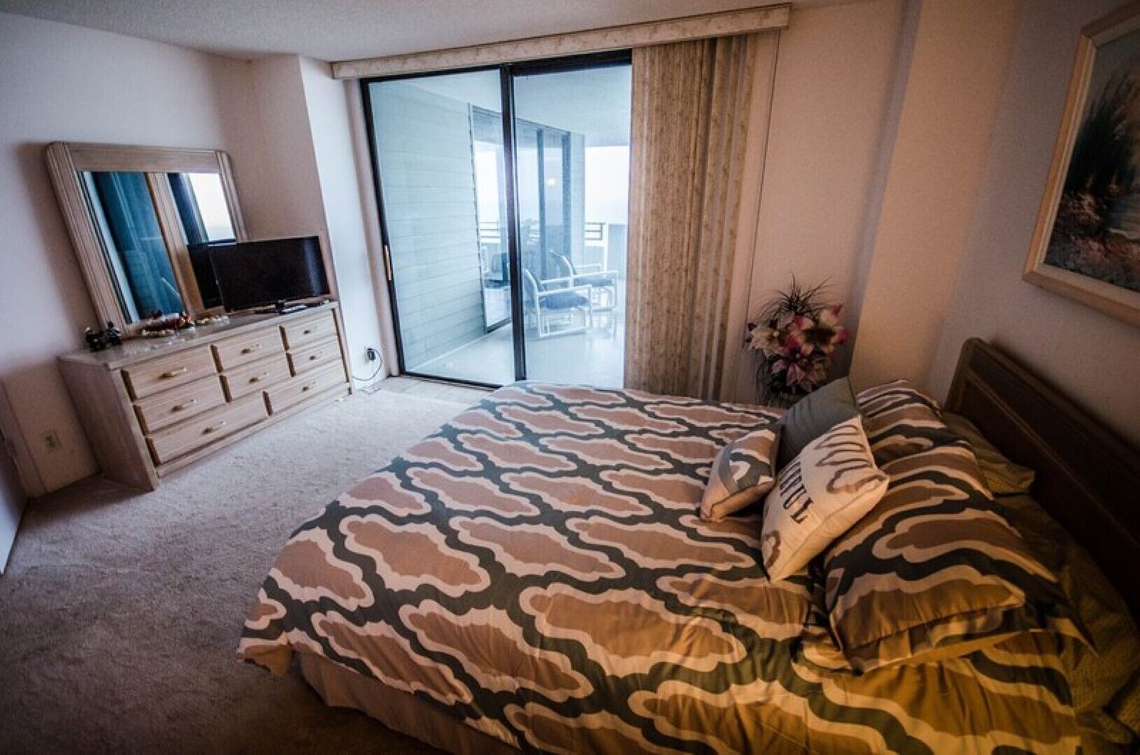 A bedroom with a bed, a dresser, a TV, a sliding glass door to a balcony, and a painting on the wall.