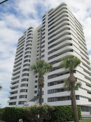 A high-rise building with multiple balconies, surrounded by palm trees and greenery.