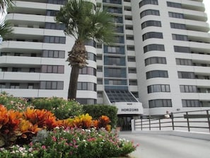 Experience the sleek, curved facade of HORIZONS, set amidst a lush garden with vibrant flowering plants and a majestic palm tree.