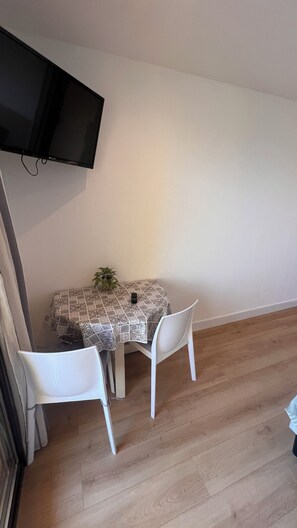 Un petit coin repas avec une table recouverte d’une nappe à motifs, deux chaises blanches, une plante en pot et un téléviseur fixé au mur.