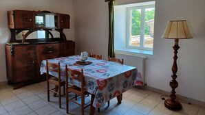 A dining area with a table set for four, a wooden cabinet, a window with curtains, and a floor lamp. - Type F2 apartment in a quiet country setting (Comps)