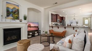Ein modernes Wohnzimmer mit Kamin, Fernseher und einer Couch. Es gibt eine Küchenzeile mit Schränken und einer Arbeitsplatte.