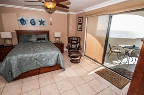 A bedroom with a bed, a chair, a ceiling fan, and a view of the ocean through a sliding glass door.