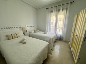 A hotel room with two beds, a nightstand, a lamp, and a window with curtains.