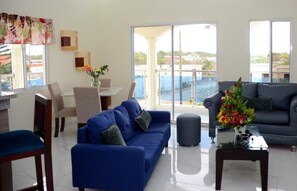 Una sala de estar con un sofá azul, un sillón negro, una mesa de centro de vidrio y un comedor con una mesa y sillas.