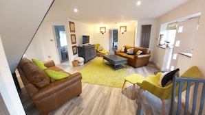 A modern living room with a brown leather sofa, a yellow armchair, and a coffee table. There is a TV on a stand and a bookshelf in the background. - Bluebell House 5 Star Luxury 4 Bdrms and hot tub (Somerton)