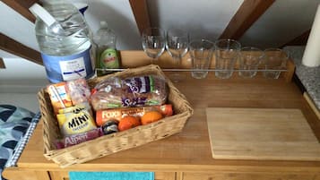Una encimera de cocina con una cesta de mimbre que contiene pan, queso y naranjas, una botella de agua, un detergente para lavar platos y varios vasos.