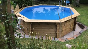 Una bañera de hidromasaje de madera rodeada por un borde de piedra, con una valla de madera y abundante vegetación al fondo.