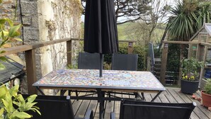 Una terraza de madera con una mesa y sillas, rodeada de plantas en macetas y un muro de piedra.