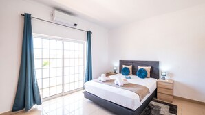 A bedroom with a bed, bedside tables, a nightstand, a window with curtains, and a wall-mounted air conditioner. - Villa with Saltwater Swimming Pool (Albufeira)