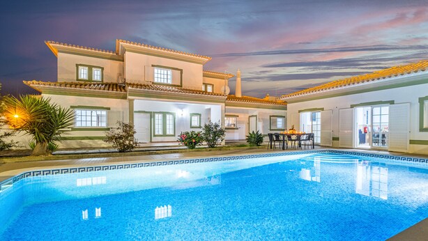 A swimming pool with a tiled deck, a patio area with seating, and a building with a tiled roof and windows. - Villa with Saltwater Swimming Pool (Albufeira)