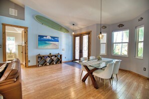 A spacious living area with a wooden floor, a dining table with chairs, a surfboard on the wall, and a beach-themed painting. - Spacious 3 Br Home w/ Pool- 1 mile walk to beach (Nokomis)