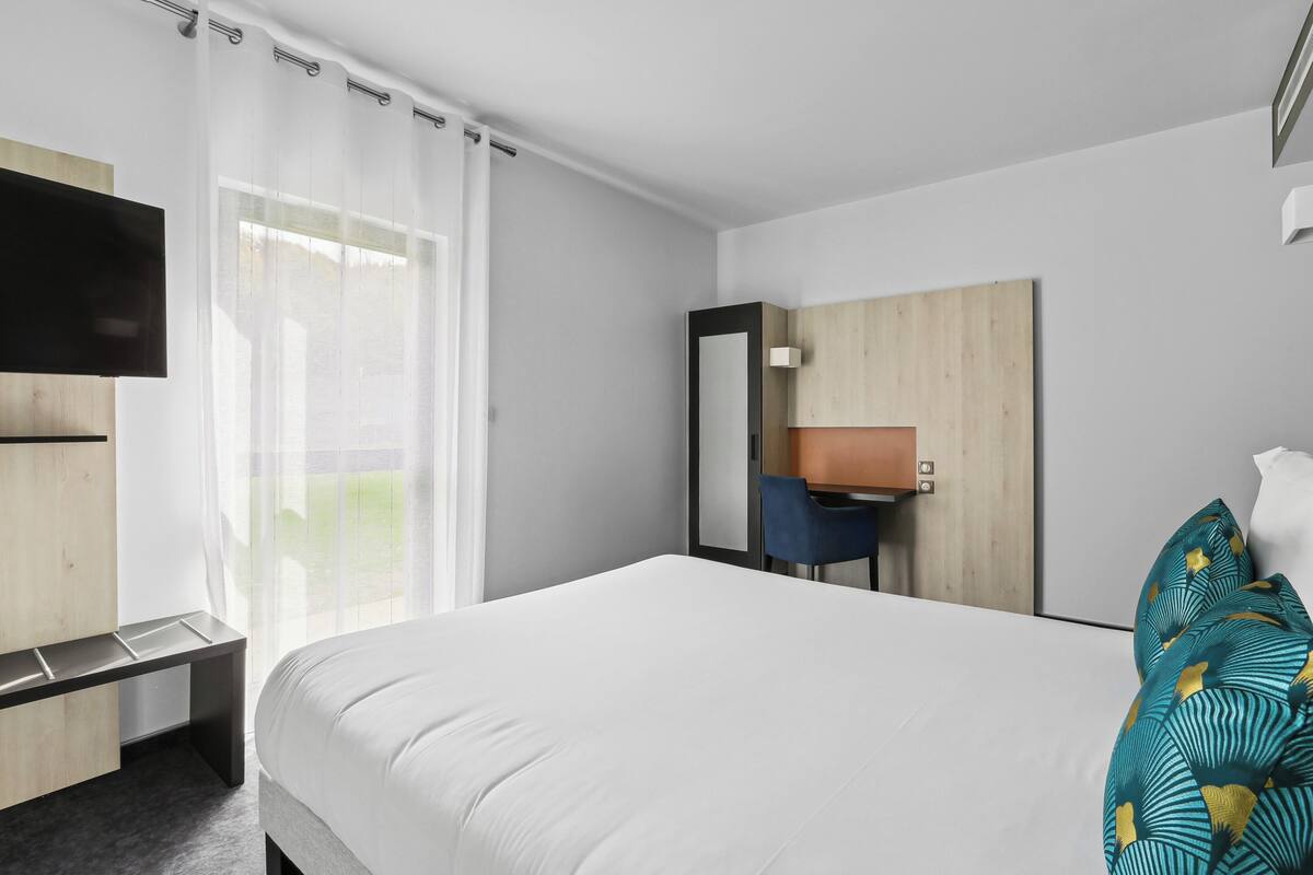 A hotel room with a bed, a desk, a chair, a TV, and a large window with curtains.
