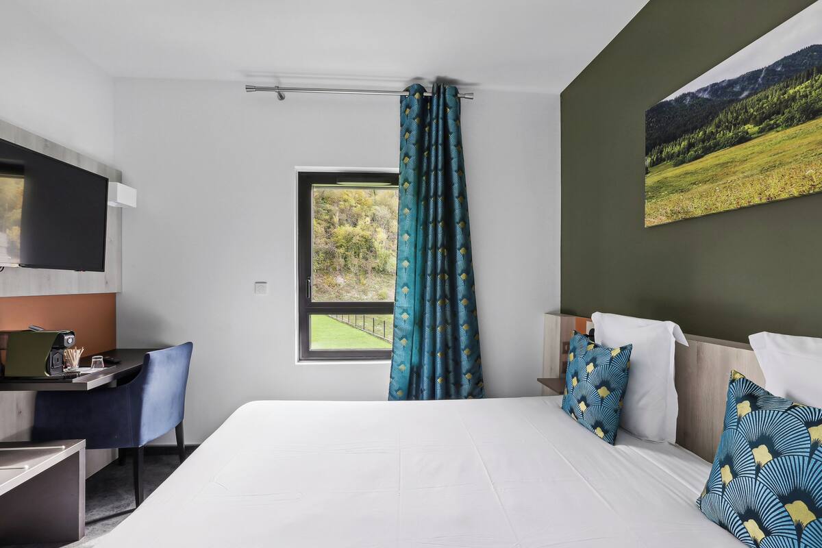 A hotel room with a bed, a desk, a chair, a window with curtains, and a painting on the wall.
