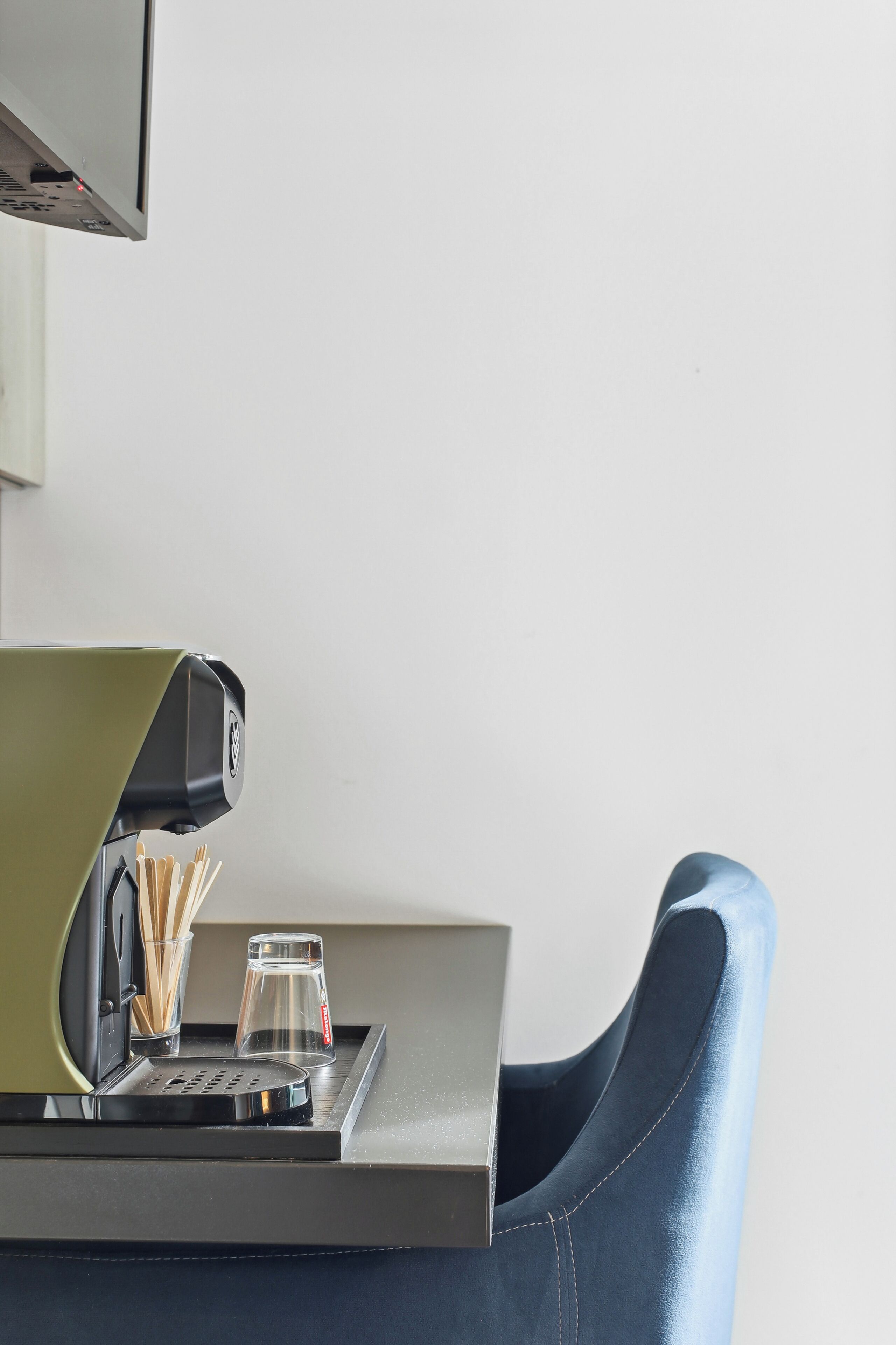 A modern coffee machine on a counter with a glass and a cup of coffee, a chair, and a TV mounted on the wall.