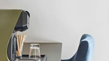 Una cafetera moderna sobre una encimera con un vaso y una taza de café, una silla y un televisor montado en la pared.