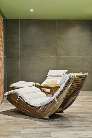 A sun lounger with a wooden frame and white cushions, positioned against a wall with large, dark tiles. - Hôtel Akena Molinges - La Brocatelle (Chassal-Molinges)